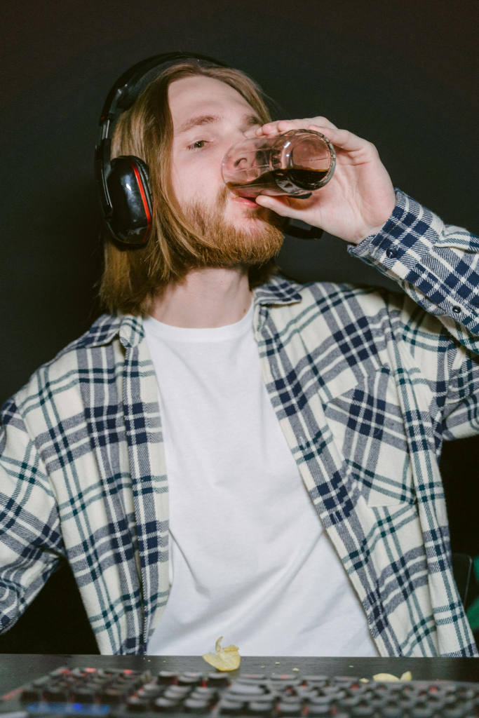 Young man gaming with headphones and plaid shirt, enjoying a drink indoors.